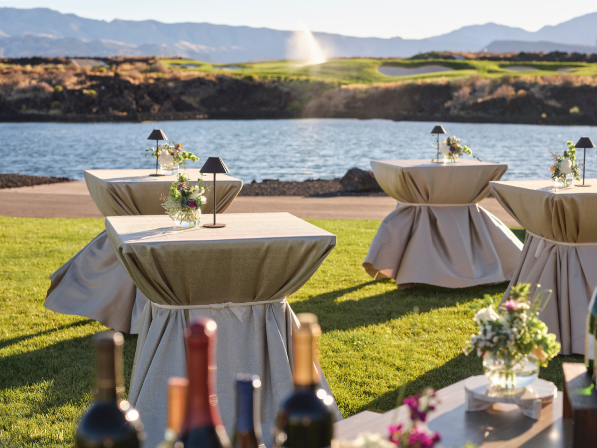 An outdoor waterfront wedding setup with draped tables, floral centerpieces, and candlelit lanterns against a sunny mountain backdrop!