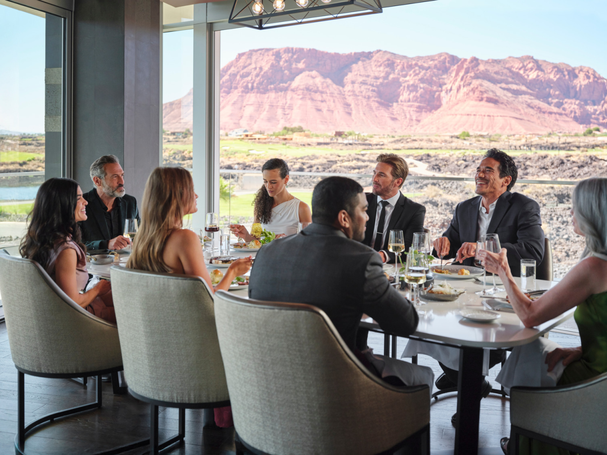 A group of people in suits dining at a restaurant with wine and a view of mountains outside the windows.