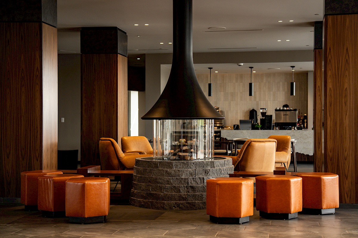 A modern hotel lobby lounge with a large stone fireplace, warm orange seating, and a coffee bar in the background.
