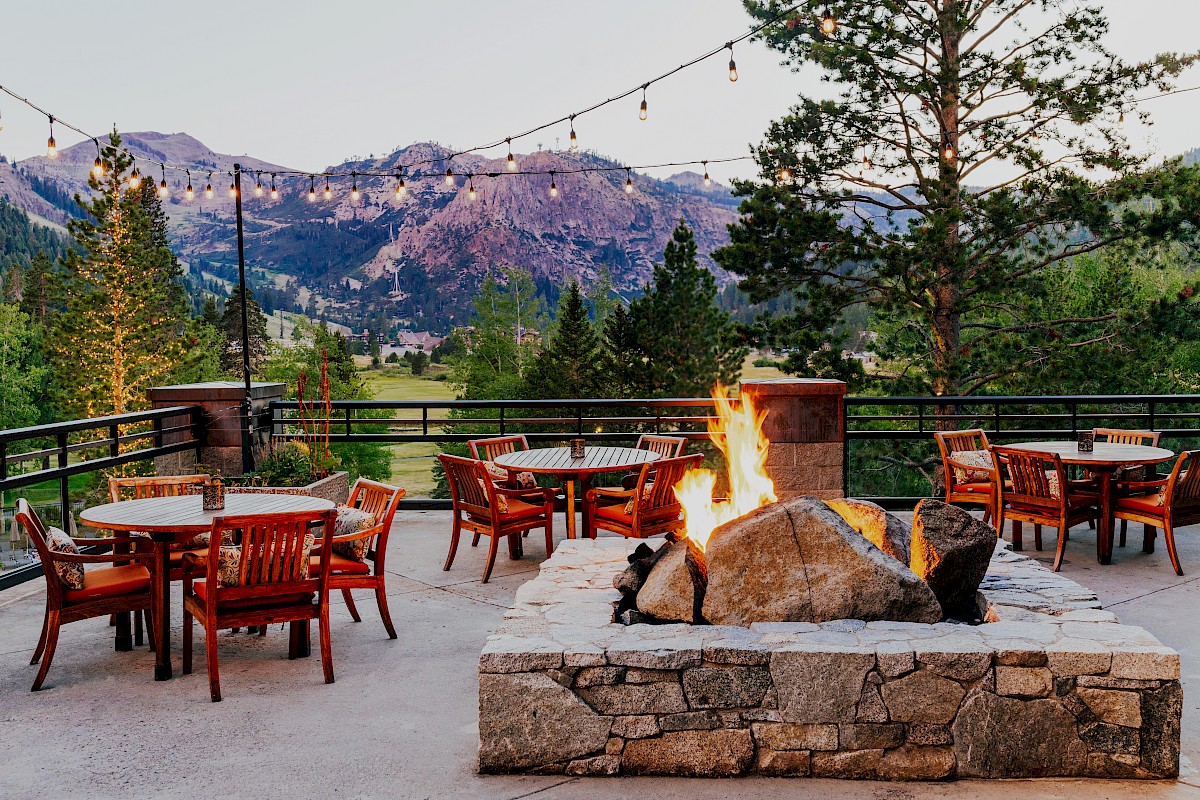 A cozy mountain patio with a roaring firepit, wooden chairs, string lights, and a scenic valley view under cloudy skies.
