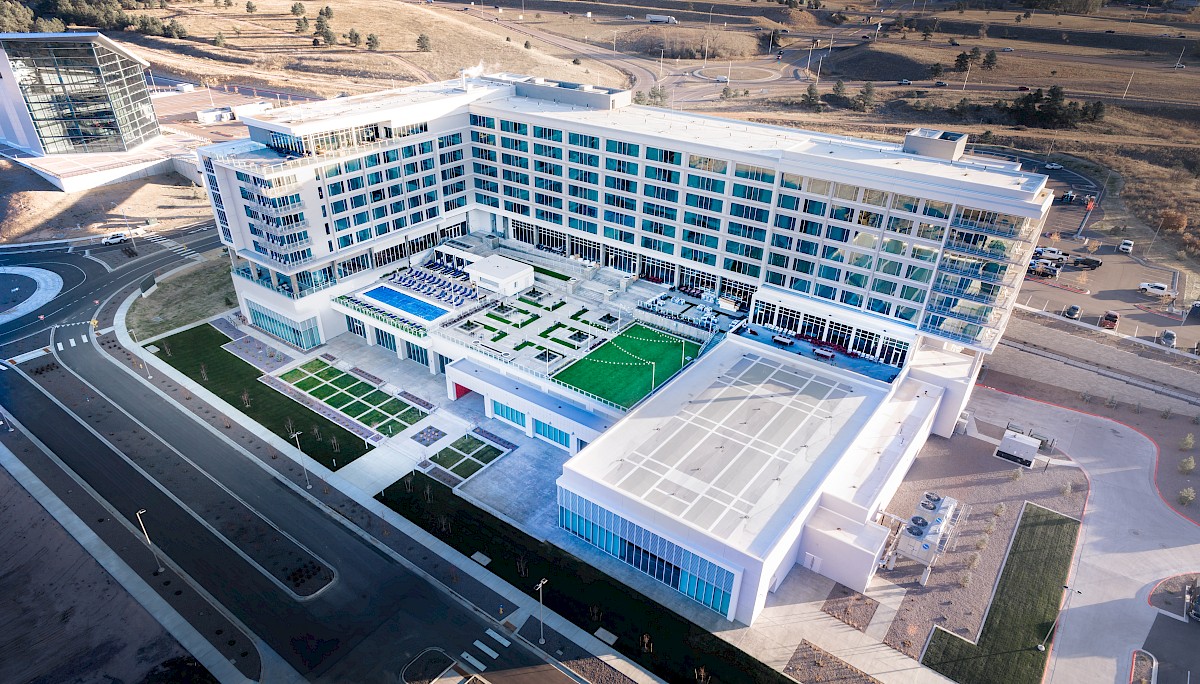 Aerial view of a modern multi-story office/campus building with a rooftop terrace, blue pool, gardens, and nearby roads.