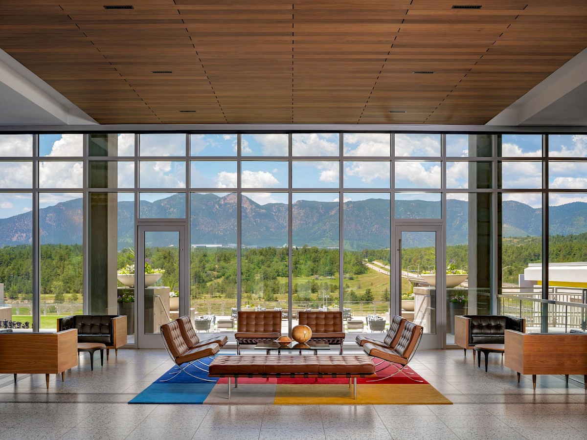 A modern lounge with leather seating, a red rug, and large windows showing green hills and mountains under a cloudy sky, facing outward.