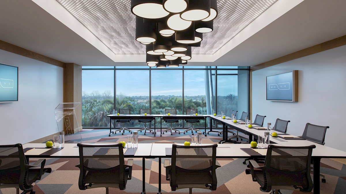 A modern office conference room with long tables, chairs, bright ceiling pendant lights, and large windows overlooking a lush landscape.