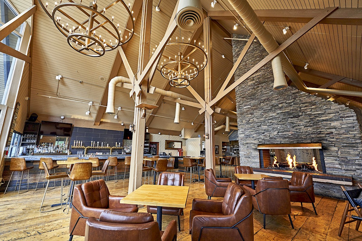 A cozy bar and lounge interior with leather seating, a large stone fireplace, and hanging globe lights above rustic wood beams.