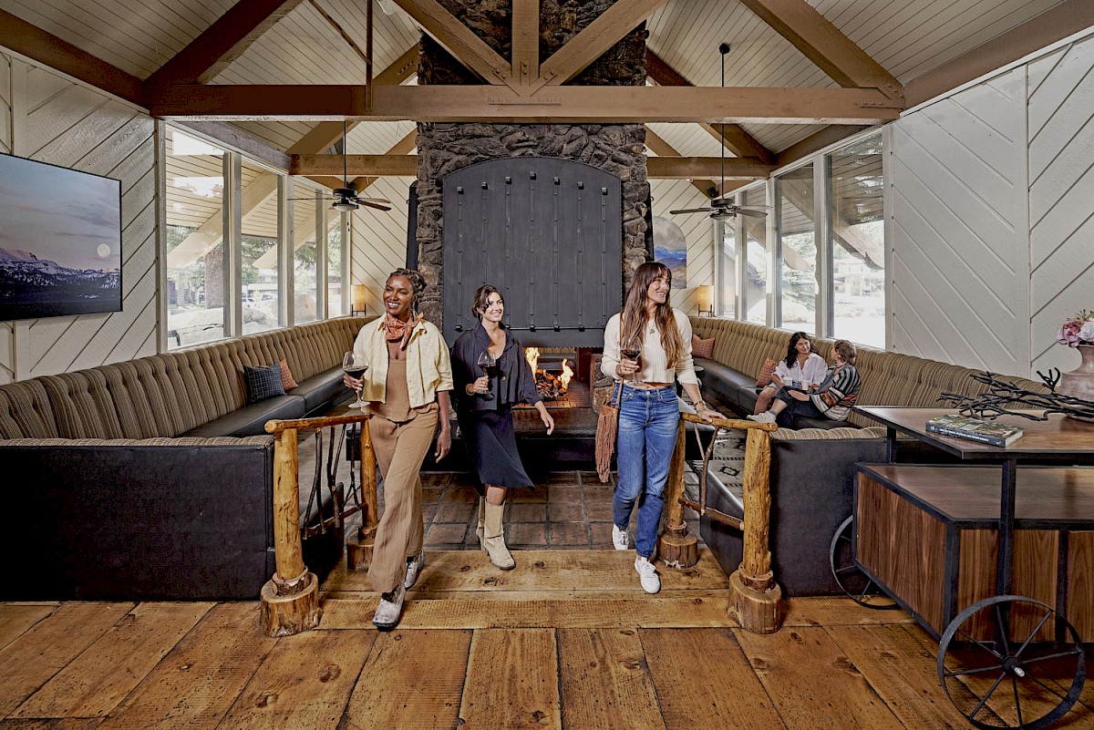 A cozy lodge living room with people standing by a crackling fireplace, holding drinks and chatting.