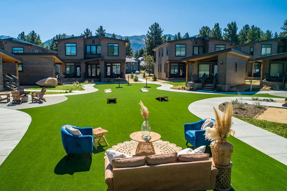 A modern apartment complex courtyard with a manicured lawn, outdoor seating, and several duplex-style buildings under a clear blue sky.