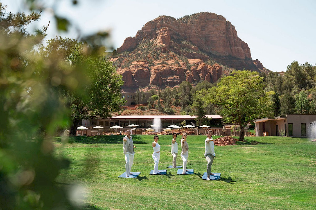 Five people are doing yoga on blue mats on a grassy lawn with a desert red rock mountain and outdoor resort buildings behind them.