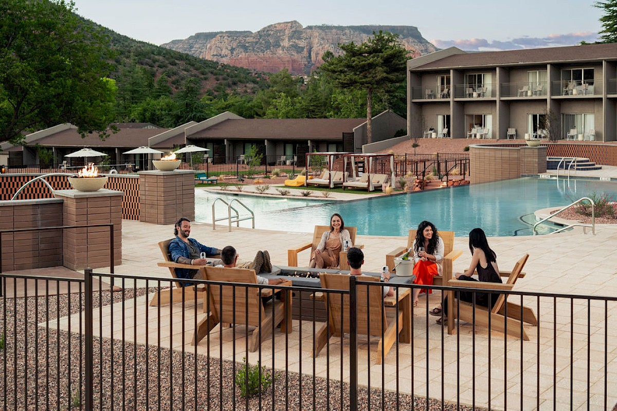 A hotel poolside scene with people relaxing on outdoor chairs under a mountain view, with a sparkling swimming pool in the background.