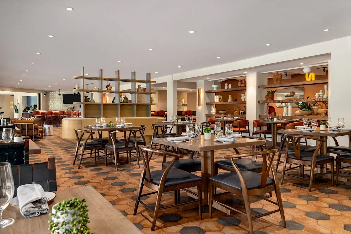 A stylish restaurant dining area with neatly set tables, warm lighting, orange seating, and an open kitchen shelf backdrop.