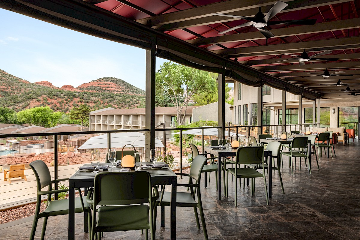 A covered outdoor dining patio with green chairs, tables set for dinner, and desert hills and trees in the background, ending.