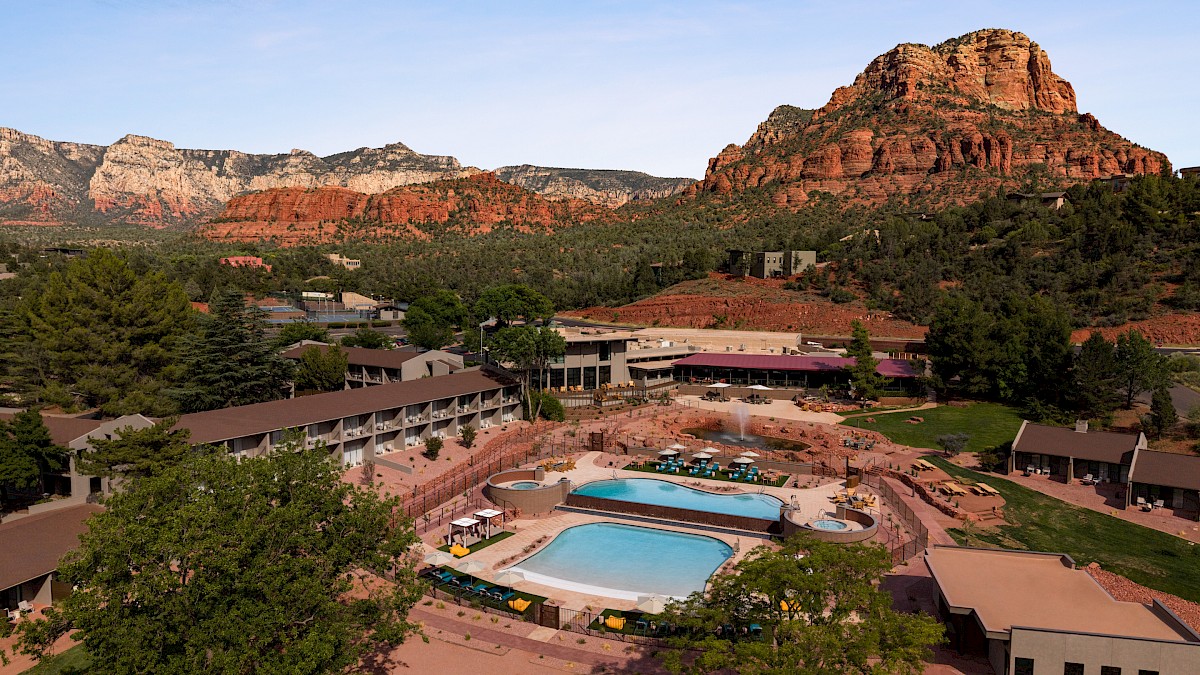 A resort hotel with two outdoor pools sits beside lush gardens, framed by dramatic red rock cliffs and mountains under a clear sky.