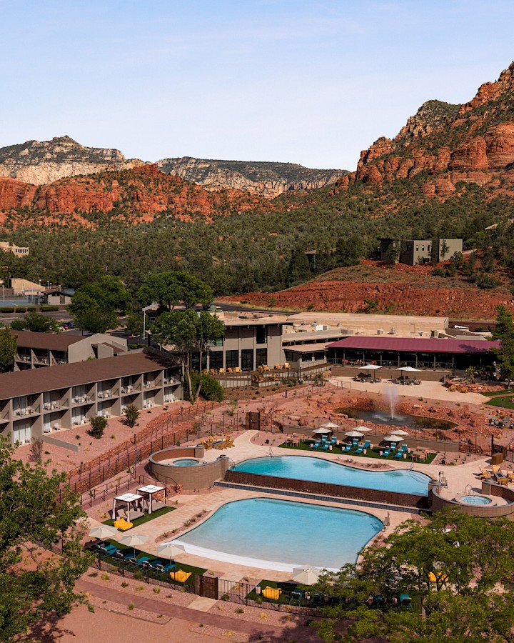 A resort hotel with two outdoor pools sits beside lush gardens, framed by dramatic red rock cliffs and mountains under a clear sky.
