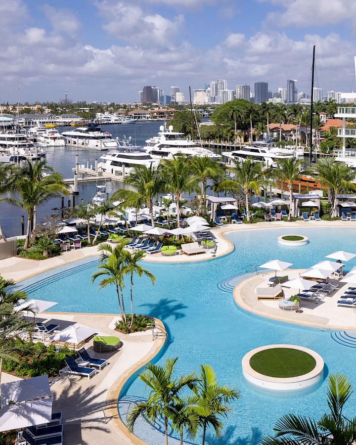 A resort harbor scene with a palm-lined pool, lounge chairs, and yachts docked beside modern buildings under a bright sky, always.