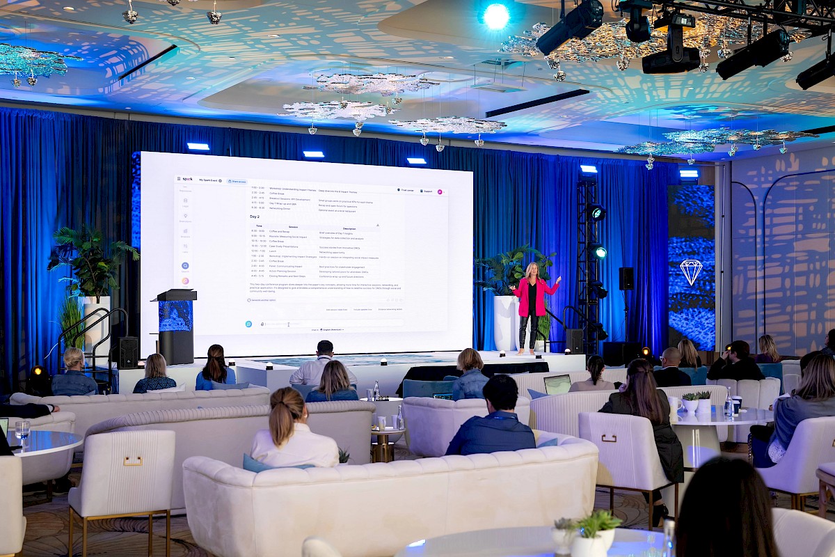 A woman presents on a conference stage with blue lighting, a large screen agenda, and an audience seated in a modern event hall.