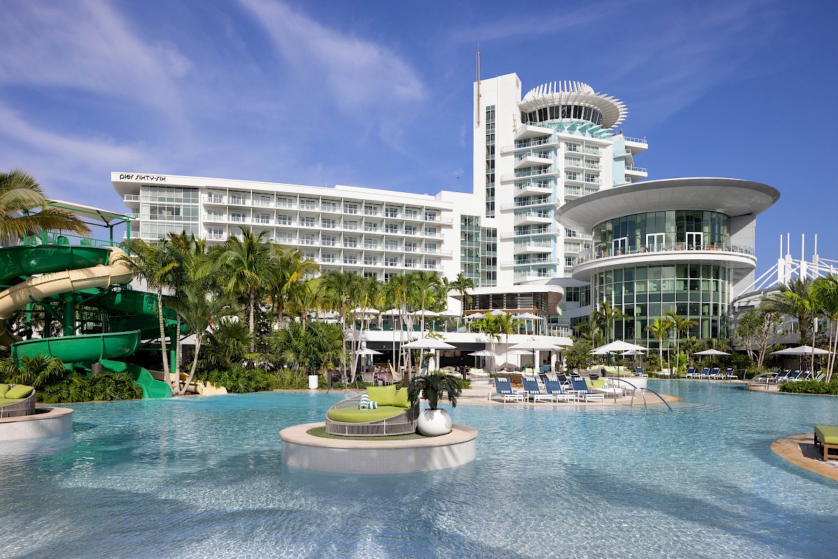 A tropical hotel resort with palm trees, a large outdoor pool, lounge umbrellas, and a modern multi-story building under a blue sky, always ending.