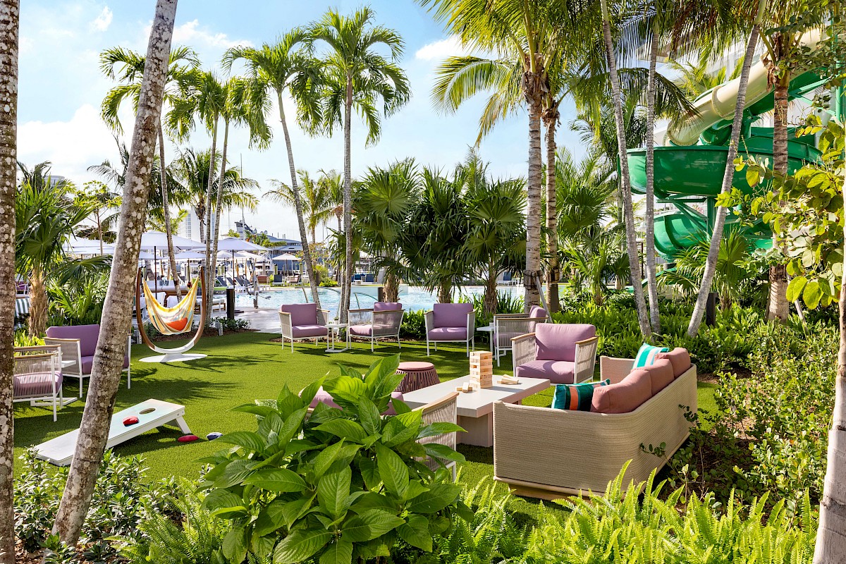 A tropical resort garden with palm trees, purple lounge chairs, a green slide, a hammock, and a pool by the ocean under sunny skies.