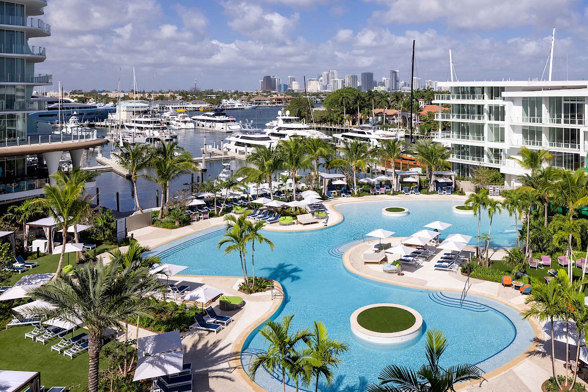 A luxury hotel marina view with sailboats, palm trees, and a large outdoor swimming pool under a cloudy sky.