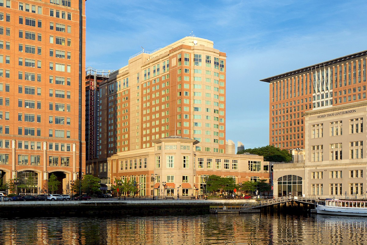 A waterfront view of downtown Boston buildings, including the World Trade Center Boston, with brick-and-glass towers reflected in the river and a clear blue sky.