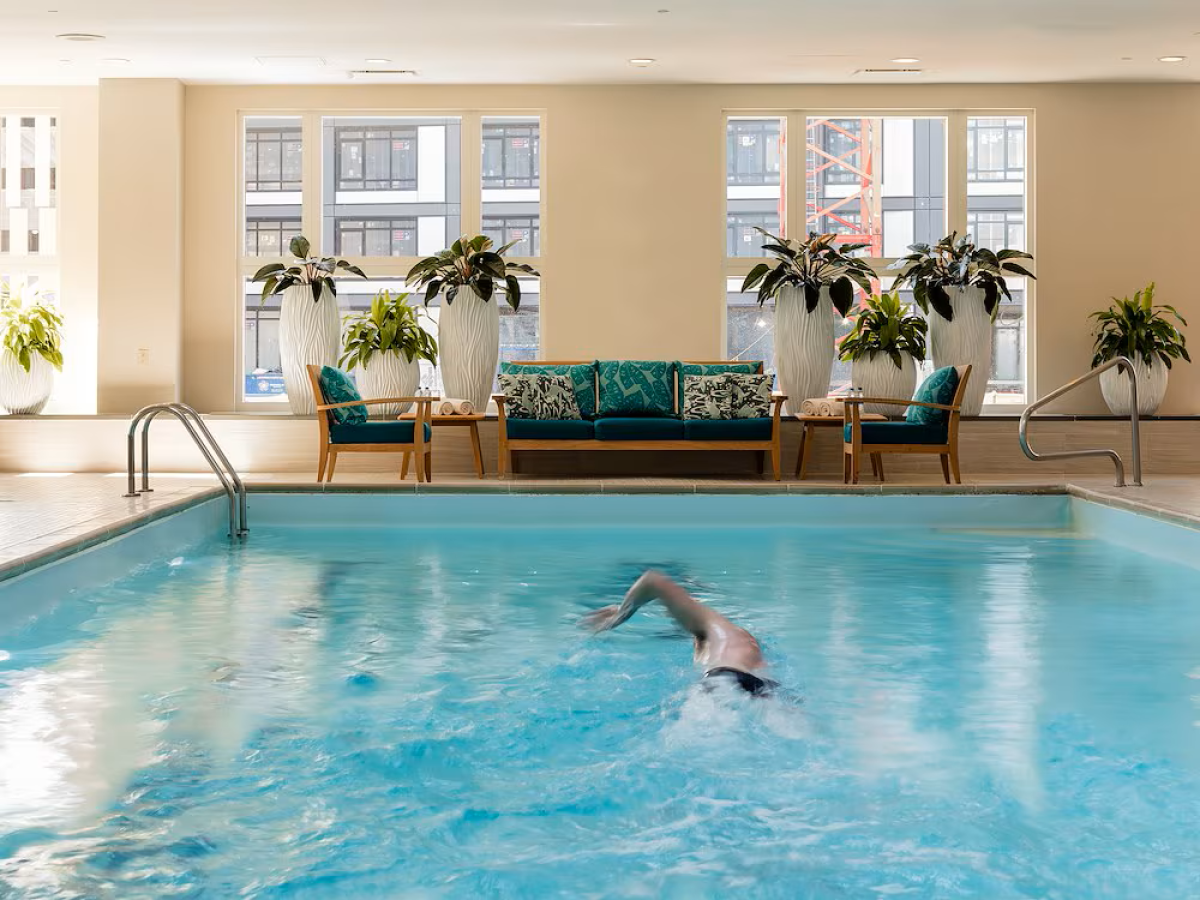 An indoor swimming pool with a swimmer and lounge seating, plants, and large windows under bright lights.