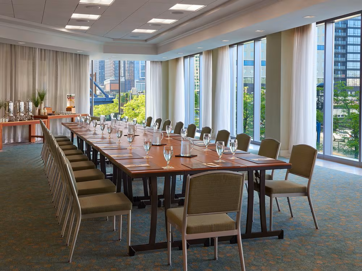 A modern hotel meeting room with a long set table, chairs, glassware, and large city view windows.