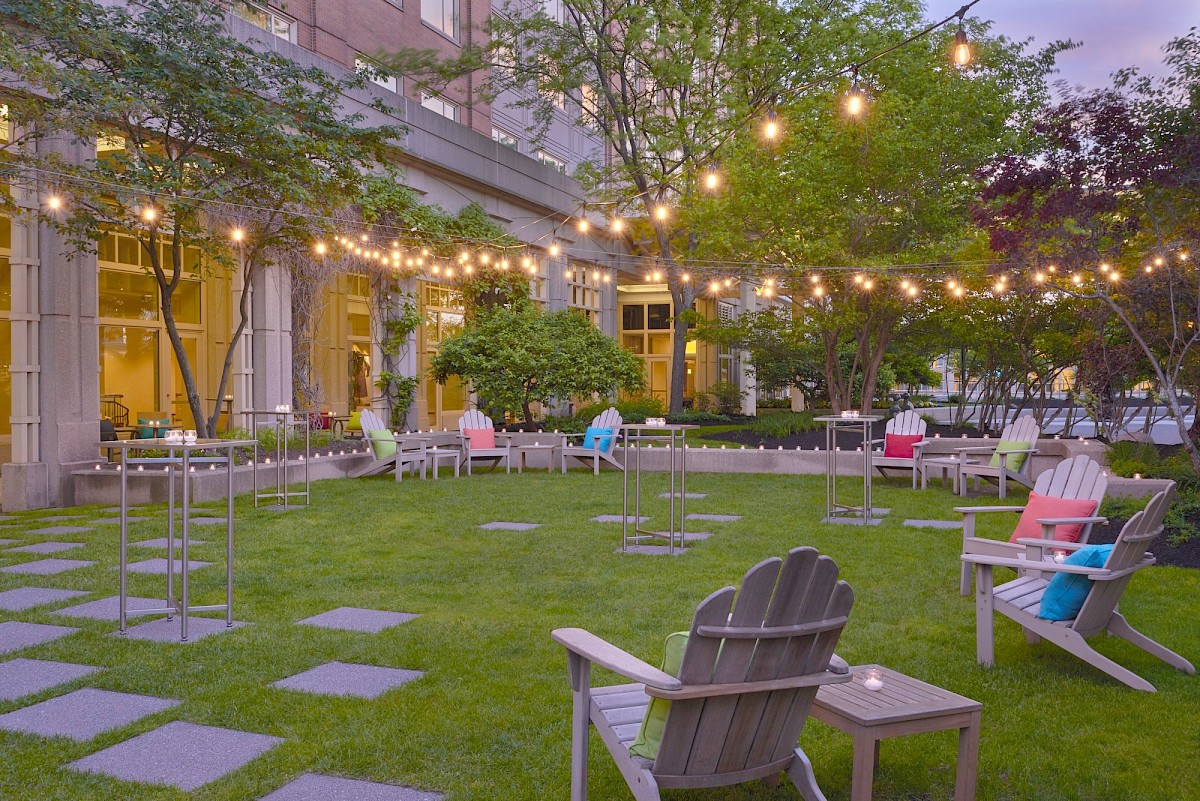 A landscaped courtyard with green lawn, colorful Adirondack chairs, tables, string lights, and a brick building—perfect for an evening gathering.
