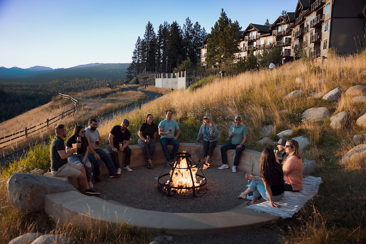 A group of friends relax by a campfire outdoors with drinks, surrounded by pine trees, grassy hills, and a mountain lodge.