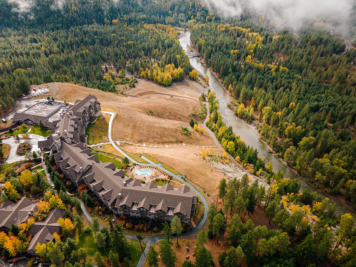 An aerial view of a mountain lodge complex beside a river valley, with forested hills and autumn foliage surrounding it.