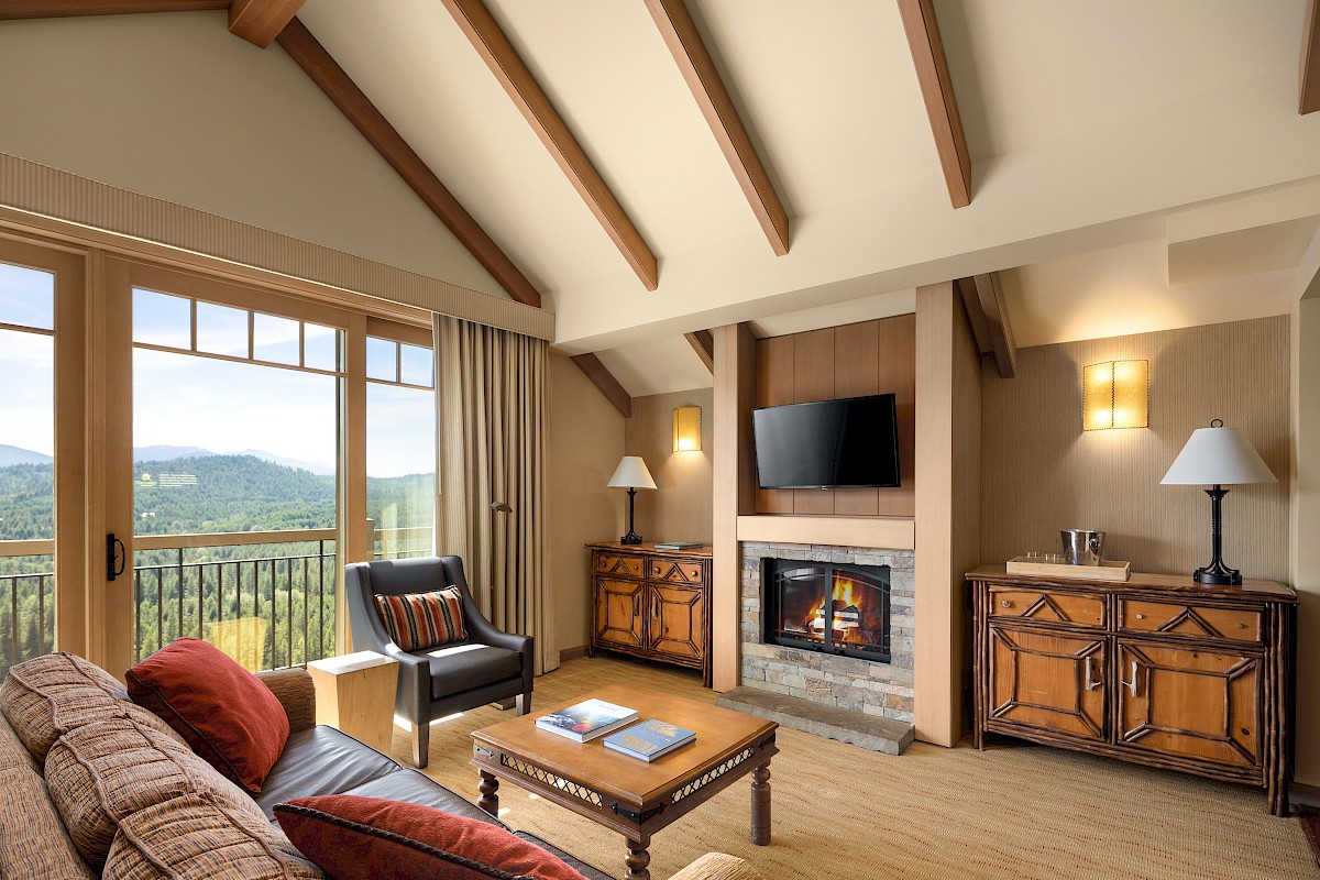 A cozy mountain living room with a fireplace, TV, lamps, wooden beams, and large windows showing a lush forest view.