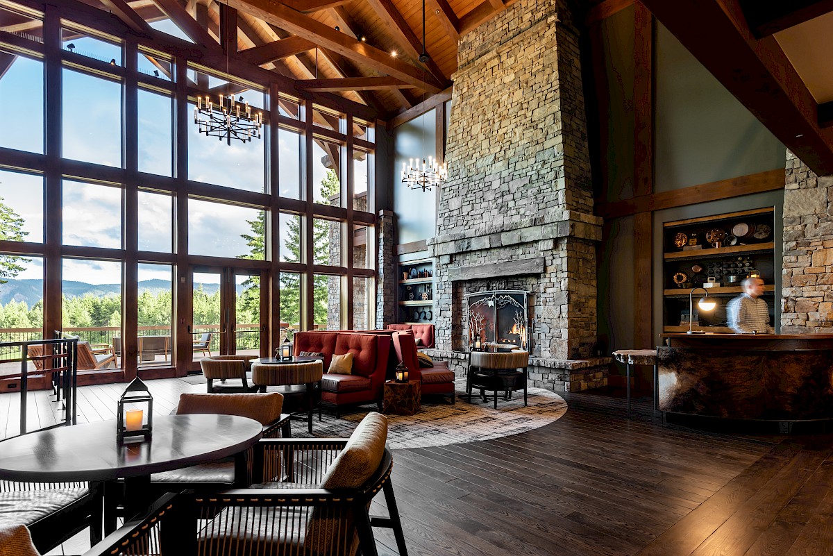 A cozy modern living room with large windows, exposed wooden beams, a stone fireplace, and warm pendant lights, ending in a relaxing mountain view.