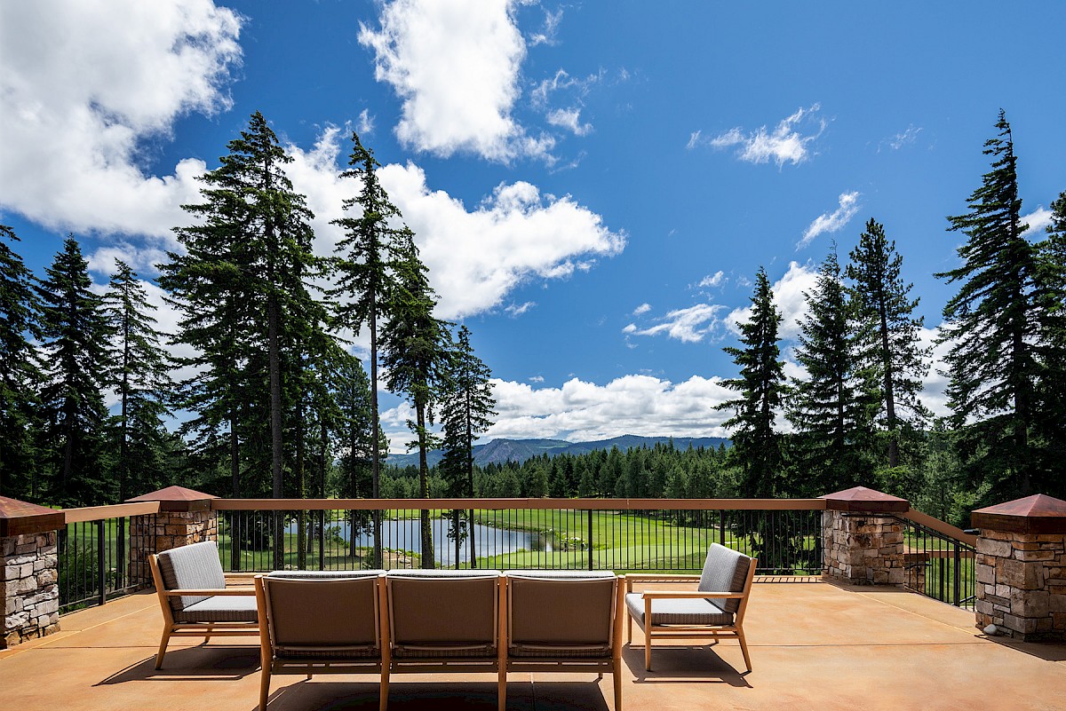 A sunny deck patio with chairs overlooking tall evergreen trees, rolling hills, and a blue sky filled with fluffy clouds.