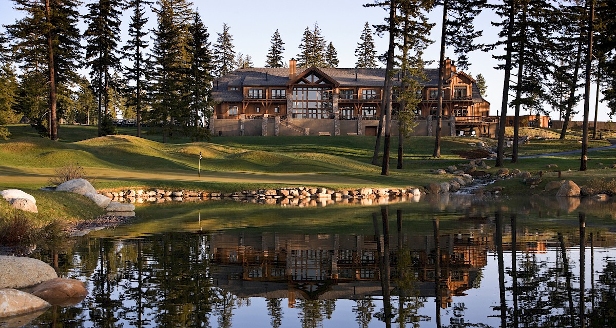 A large wooden lodge with balconies sits beside a calm pond, surrounded by tall evergreen trees, rocks, and a stone-lined shoreline at sunset.