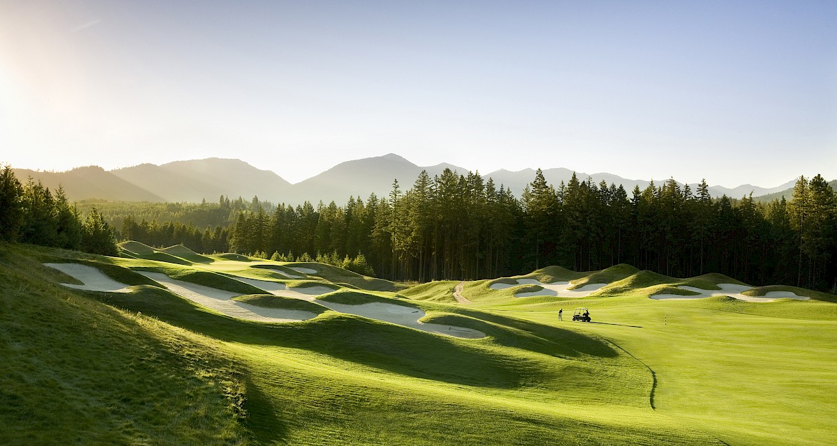 A sunny golf course with rolling fairways, sand bunkers, forest trees, and distant mountains with a golf cart on the green.
