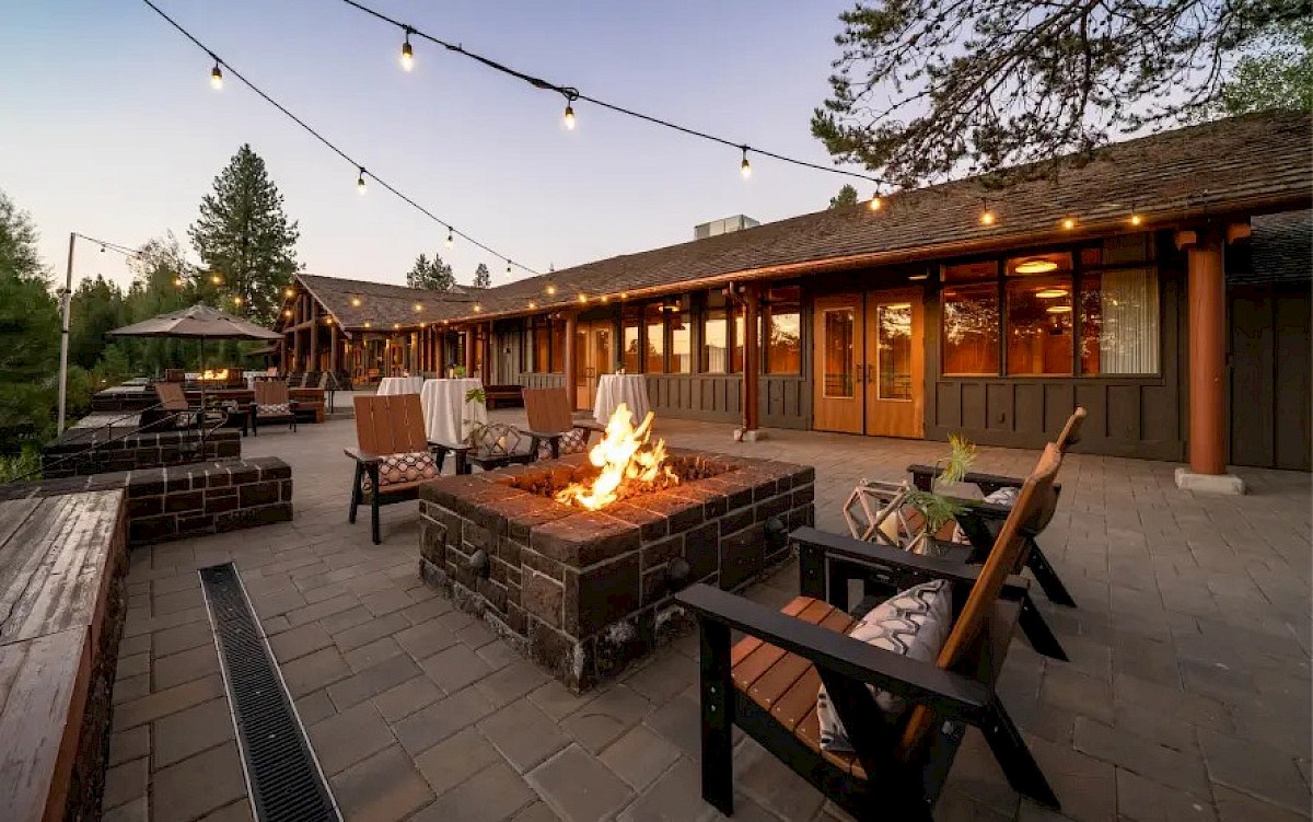 An outdoor patio at dusk with string lights, a stone fire pit, and patio seating and umbrellas around it.