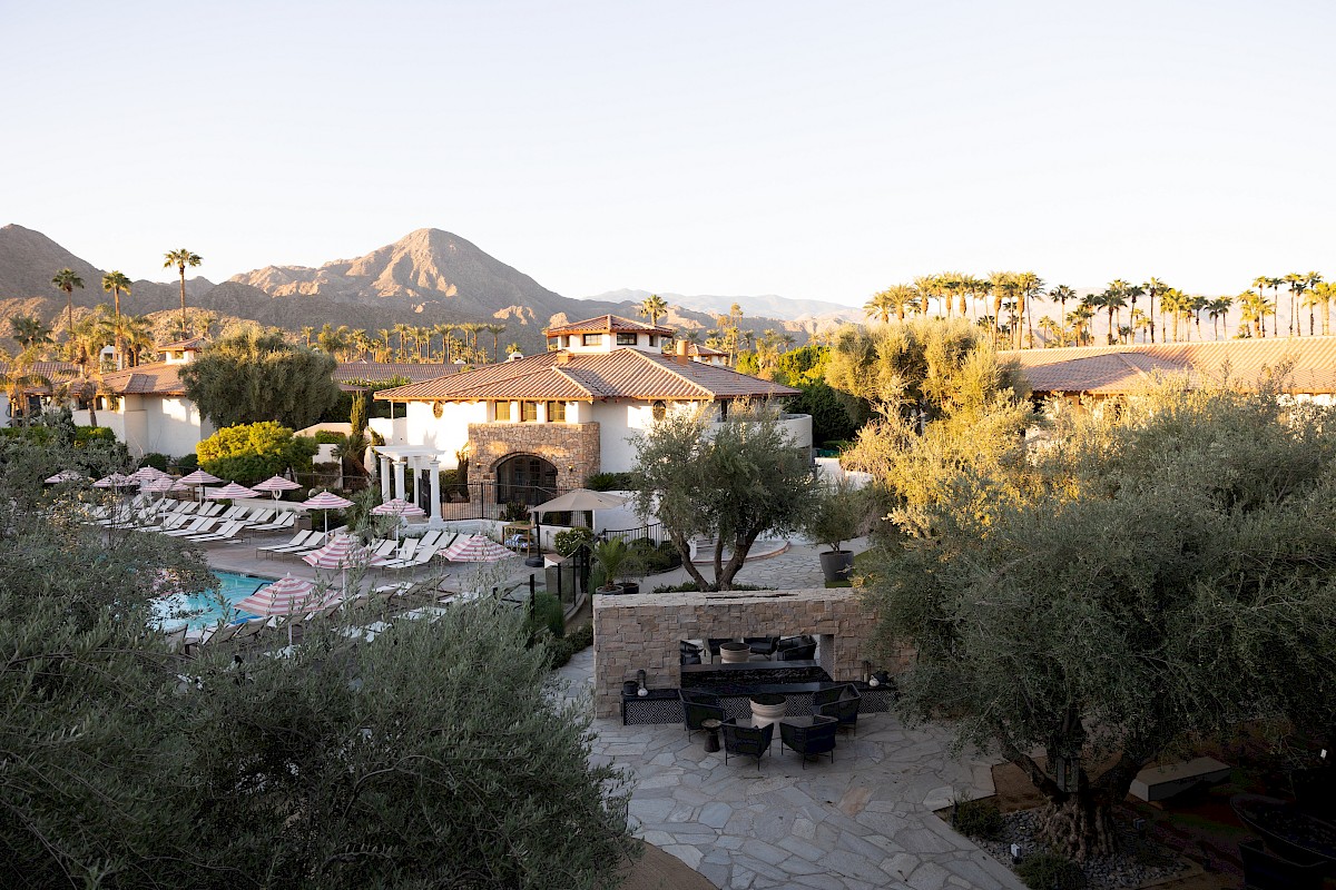 A luxury resort courtyard with a swimming pool, lounge umbrellas, white villas, palm trees, and mountains at sunset.