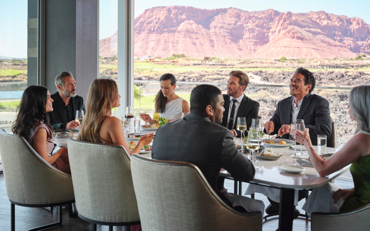 A group of people in formal attire dine together at a restaurant table with mountain scenery visible through large windows.