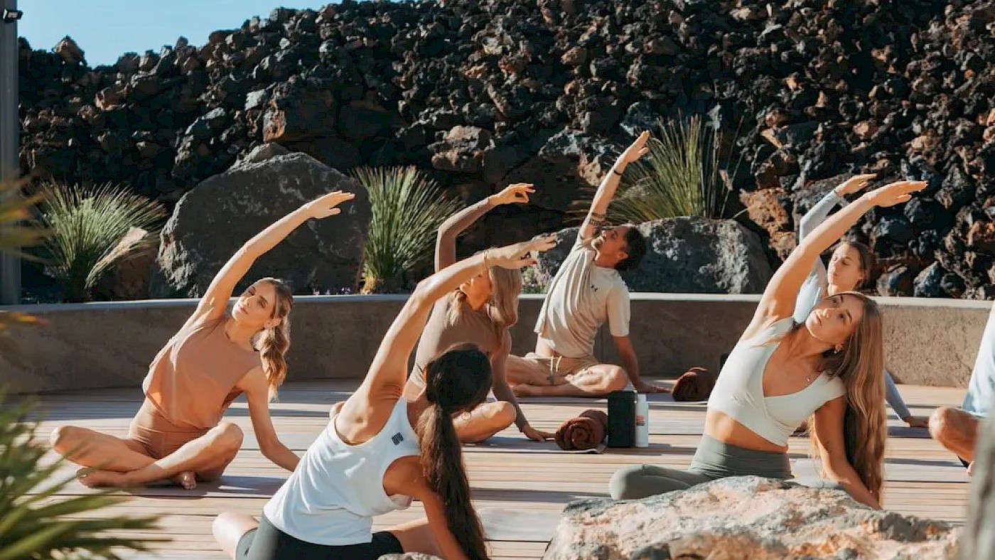 A group of women practice yoga poses on an outdoor deck, stretching their arms toward the sky against a rocky hillside backdrop.